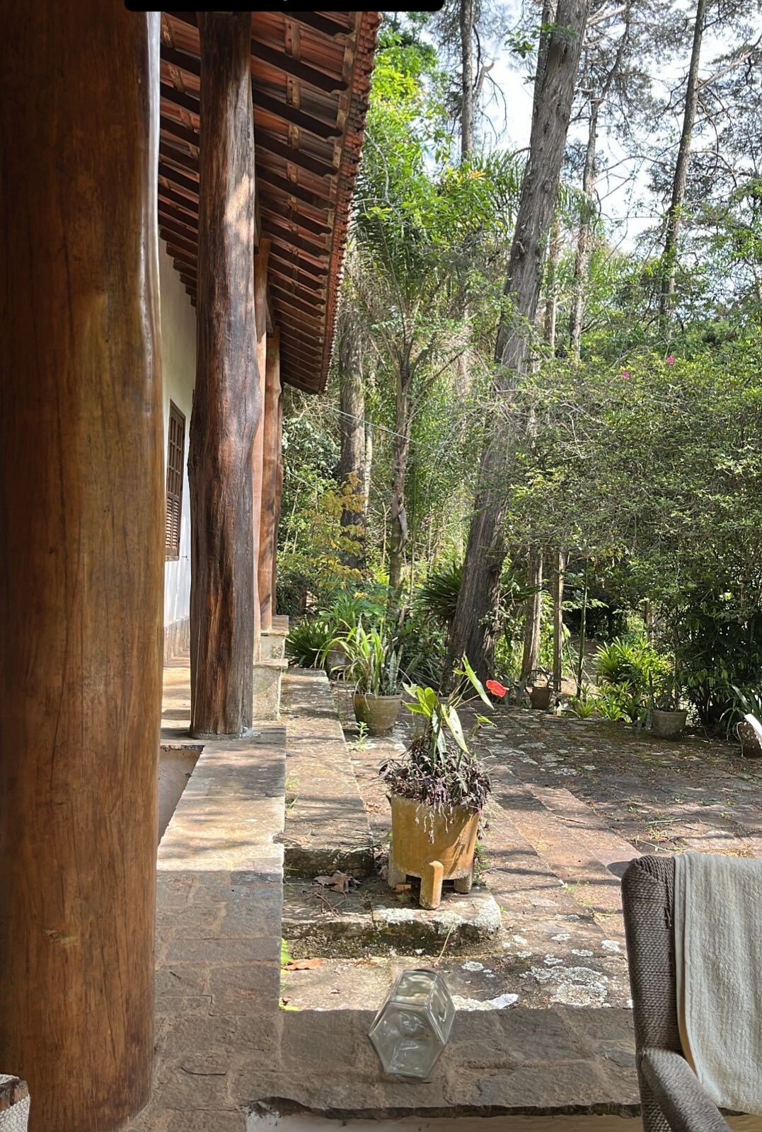 A serene outdoor area is framed by sturdy wooden columns, showcasing natural stone flooring. Lush greenery, including various plants and trees, creates a tranquil environment. A simple container with plants adds a touch of nature, inviting relaxation and appreciation of the surrounding landscape.
