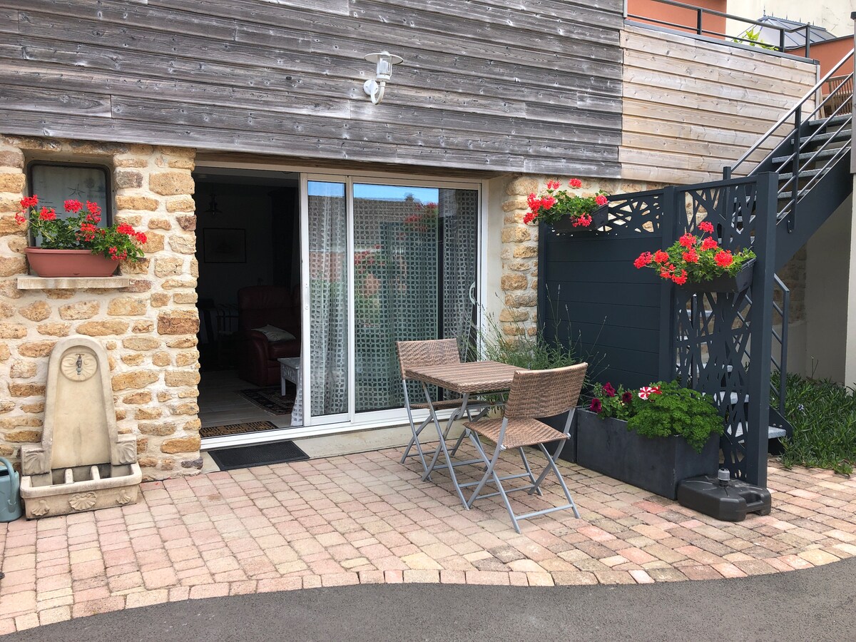 An outdoor terrace is presented with a small bistro table and two wicker chairs, surrounded by planters filled with vibrant flowers. The entrance features sliding glass doors framed by decorative curtains, leading into the cozy interior space visible in the background.