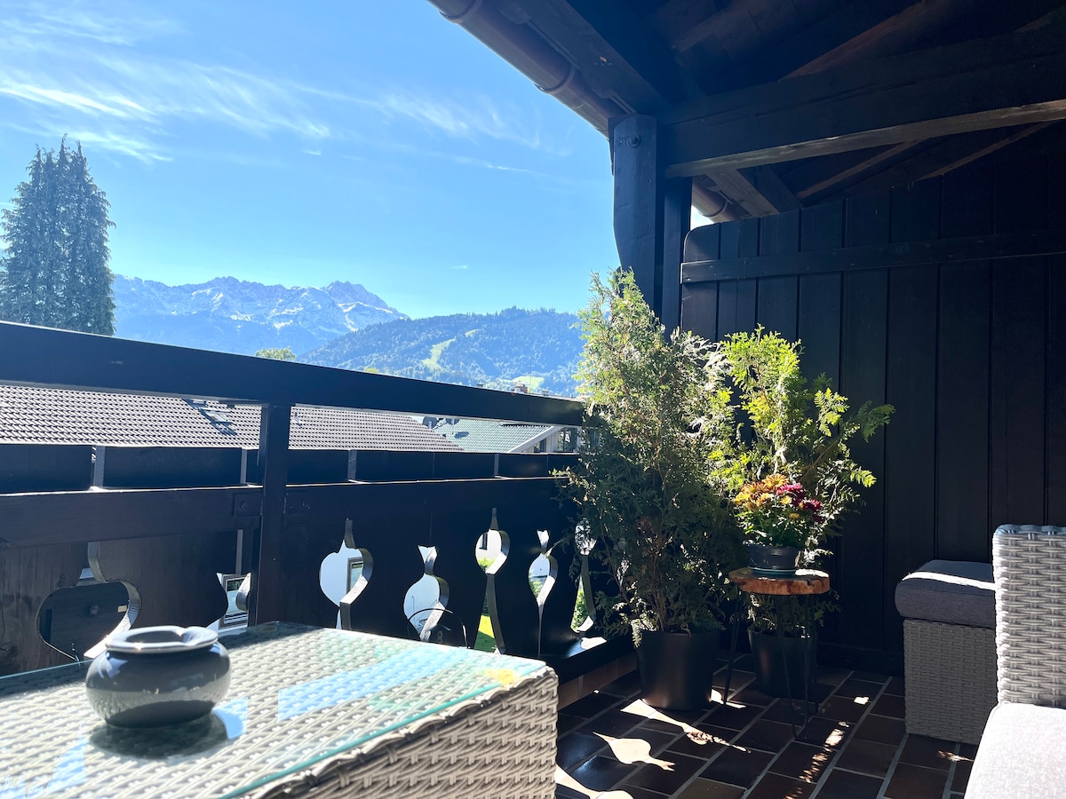 A loggia features a stylish lounge set and views of the surrounding mountains. Lush greenery is positioned in pots, enhancing the serene setting. The space is sheltered by a wooden overhang, providing a comfortable outdoor experience.