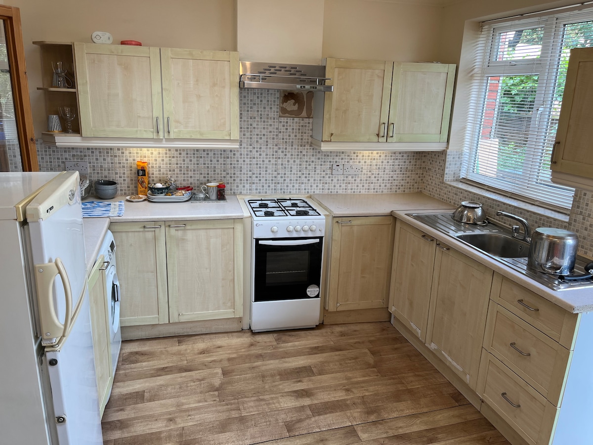 A well-equipped kitchen features light wood cabinetry and modern appliances. A gas stove is positioned beside a stainless steel sink. Natural light enters through a window adorned with blinds, illuminating the counter space. The floor displays a warm, wood-like finish, contributing to the room's cozy ambiance.