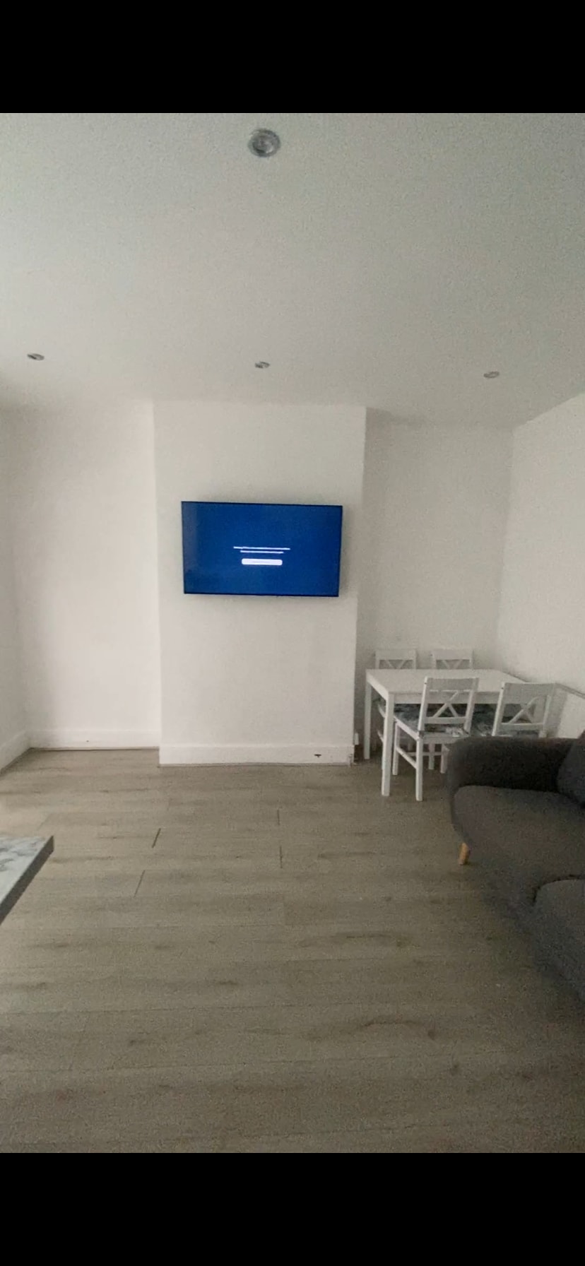 A bright open-plan living area contains a comfortable gray sofa and a dining table with four white chairs. A wall-mounted television is positioned above a smooth surface, and natural light is subtly enhancing the minimalist design of the space.