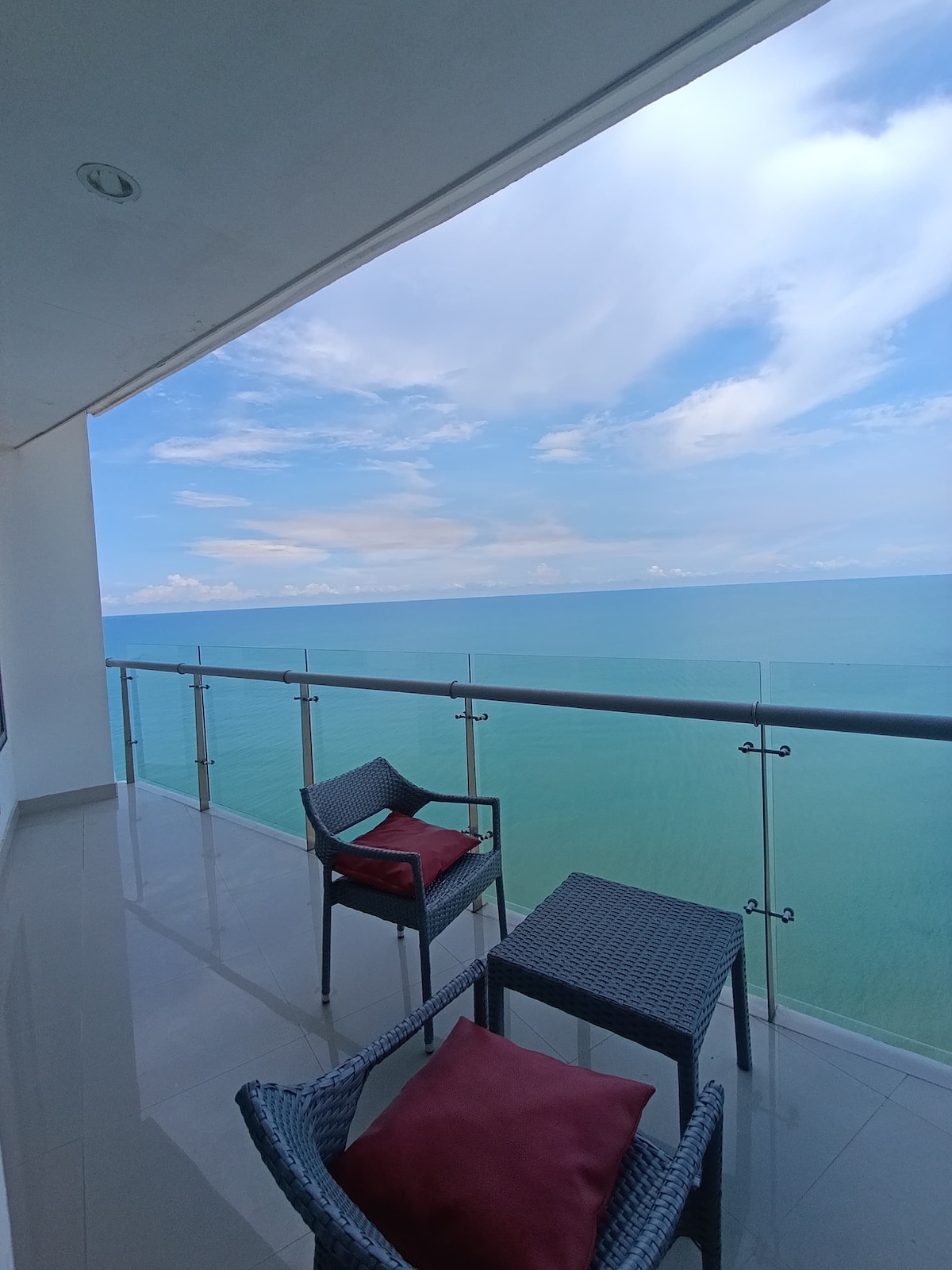 A spacious terrace is presented, featuring outdoor furniture including two chairs with red cushions and a small table. A stunning view of the calm ocean extends to the horizon, with soft blue skies and wispy clouds enhancing the serene atmosphere.