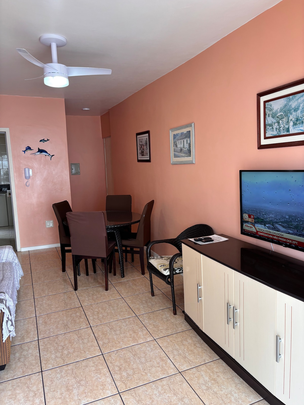 A dining area features a glossy table surrounded by four brown chairs, with a dark wooden sideboard nearby. A television is mounted on the wall, displaying a scenic image. Light-colored tiles cover the floor, and the walls are painted in a soft peach hue.