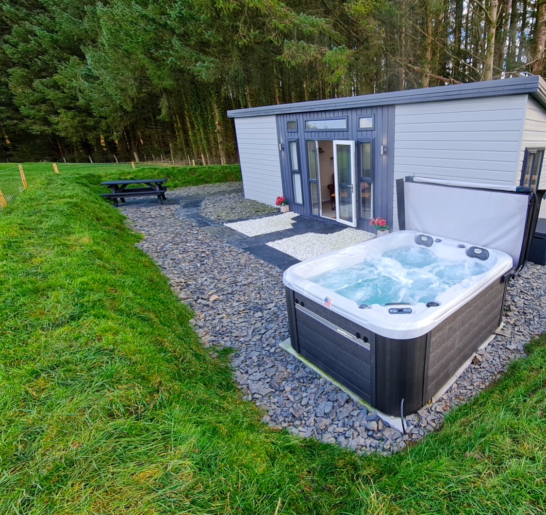 A hot tub is positioned on a gravel area adjacent to a lodge, surrounded by greenery. The lodge features large glass doors, allowing for natural light. Outdoor seating is available nearby at a picnic table, providing a casual space for relaxation.