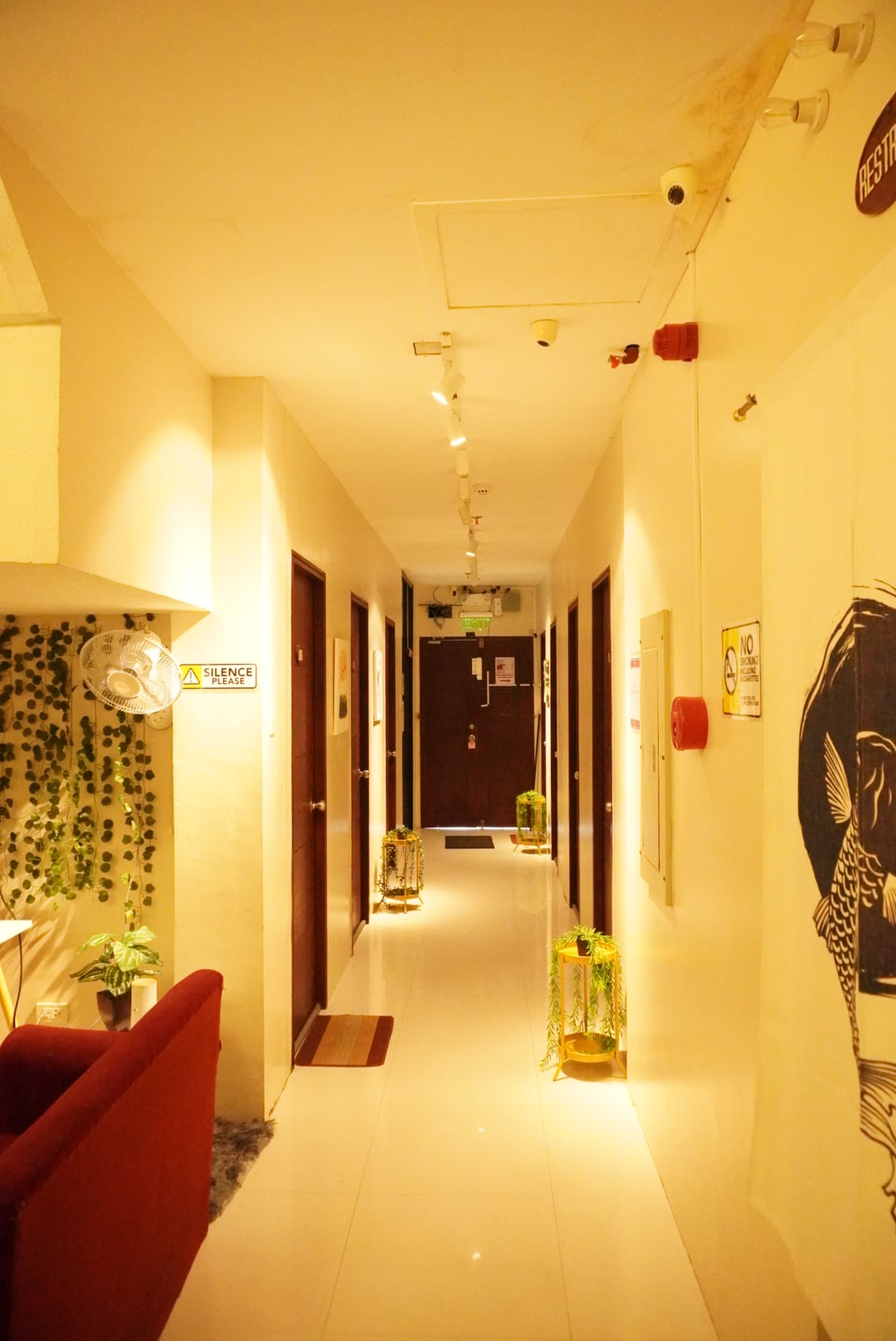 A narrow hallway is illuminated with soft lighting, featuring multiple closed doors on both sides. Potted plants are positioned along the floor, contributing to a fresh ambiance. A small seating area with a Scandinavian chair is visible at the end of the corridor.