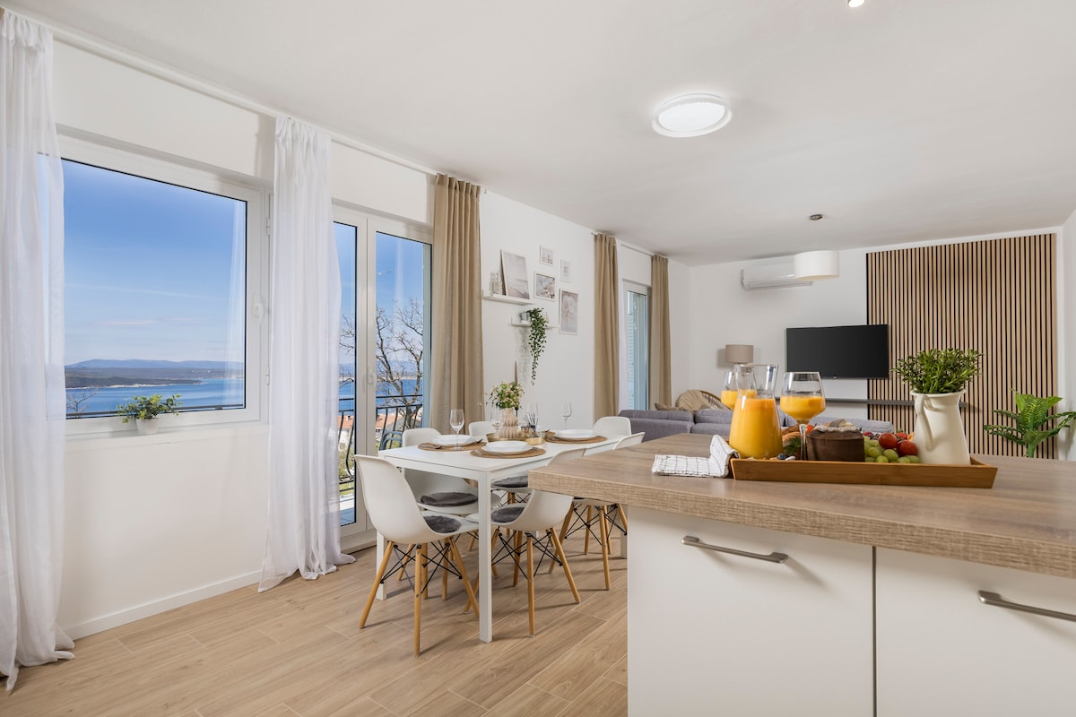 A light-filled open living space features large windows offering a scenic view. A dining table with four chairs is set for a meal, while a comfortable bed is visible in the background. Refreshing fruits and drinks are displayed on a wooden tray in the kitchen area.