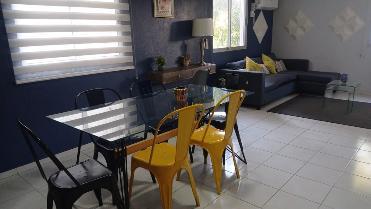 A dining area features a glass table surrounded by four black chairs and two yellow chairs, offering a lively contrast. A comfortable sectional sofa is positioned in the corner, complemented by decorative pillows. Light filters through window treatments, illuminating the space.