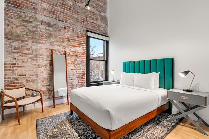 Queen bedroom with velvet headboard, exposed brick accent, leaning mirror, original window.