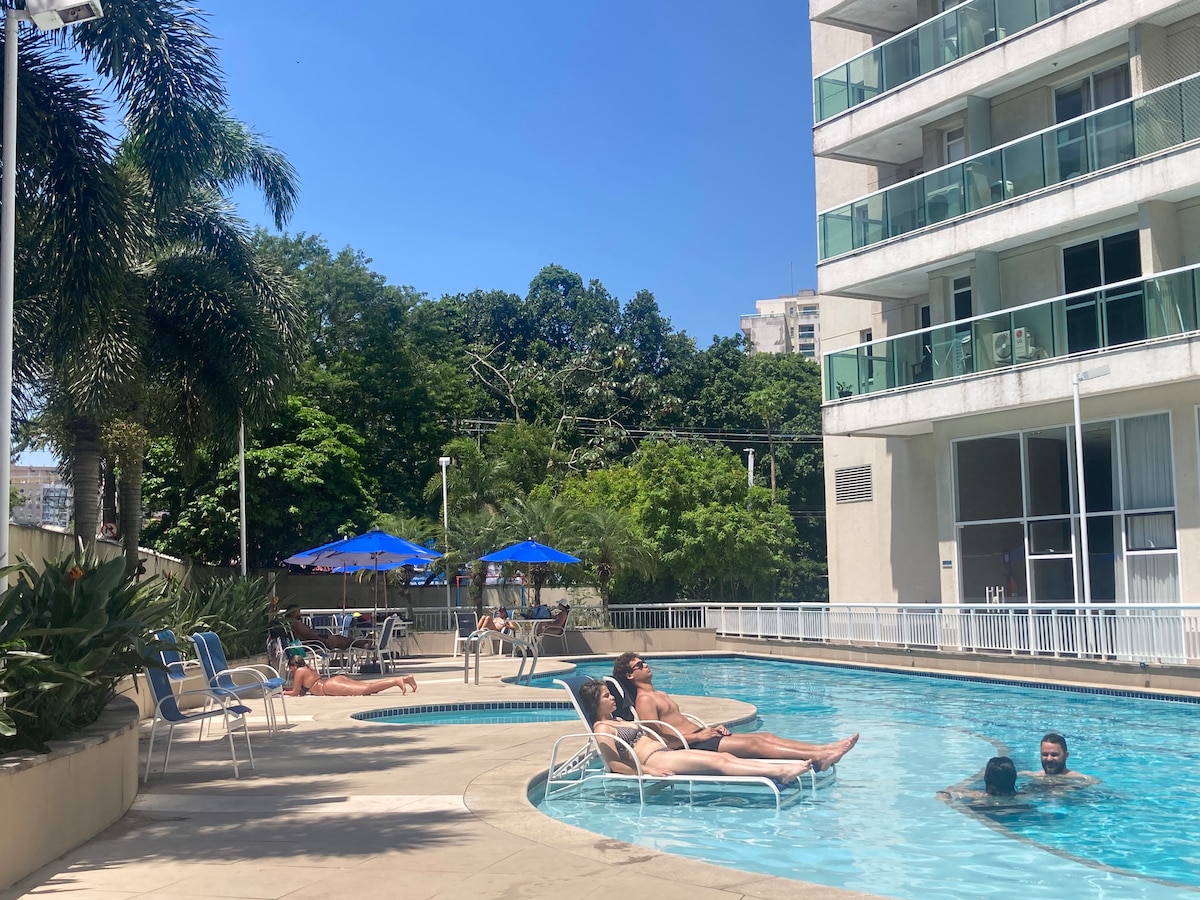 The pool area features a curved shape with lounge chairs positioned around it. Sun umbrellas provide shade, and there are several trees in the background. The design promotes relaxation, with guests enjoying the warm weather by the water.