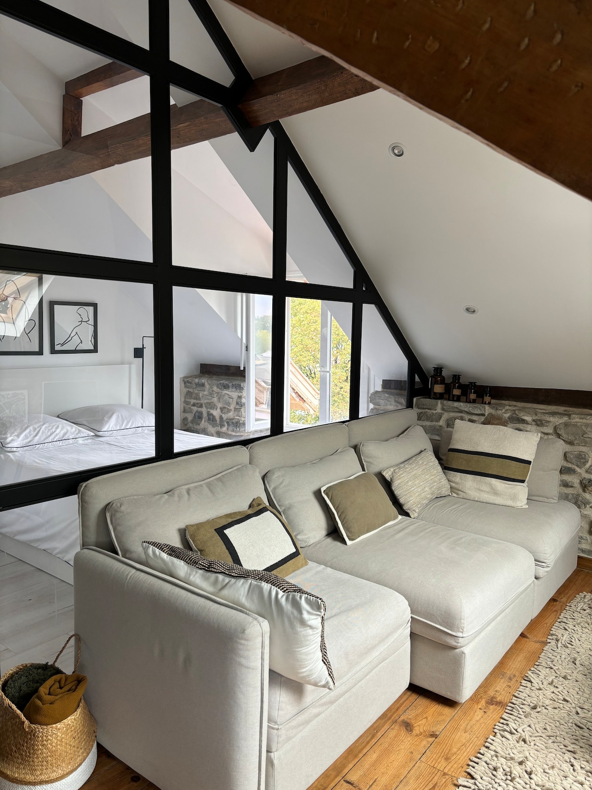 A light-filled living area features a comfortable sectional sofa adorned with decorative pillows. The room showcases a blend of modern and rustic elements, with exposed wooden beams and a stone wall. A separate sleeping area is visible through large glass panels.