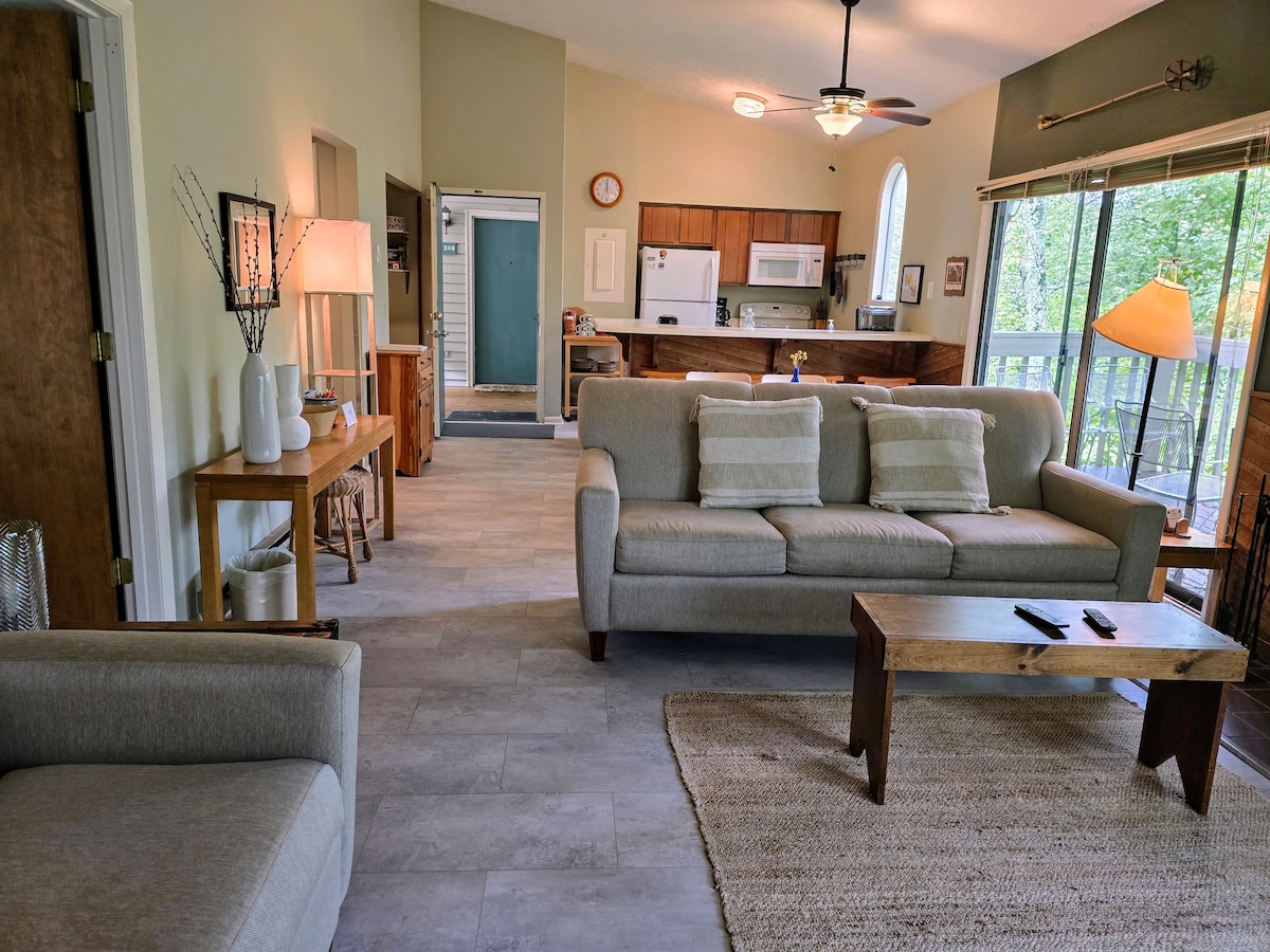 A spacious living area displays comfortable seating, including a neutral-toned sofa and armchair. A wooden coffee table is centered in the space. The kitchen is visible in the background, featuring wood cabinetry and modern appliances. Light enters through large glass doors, enhancing the inviting atmosphere.