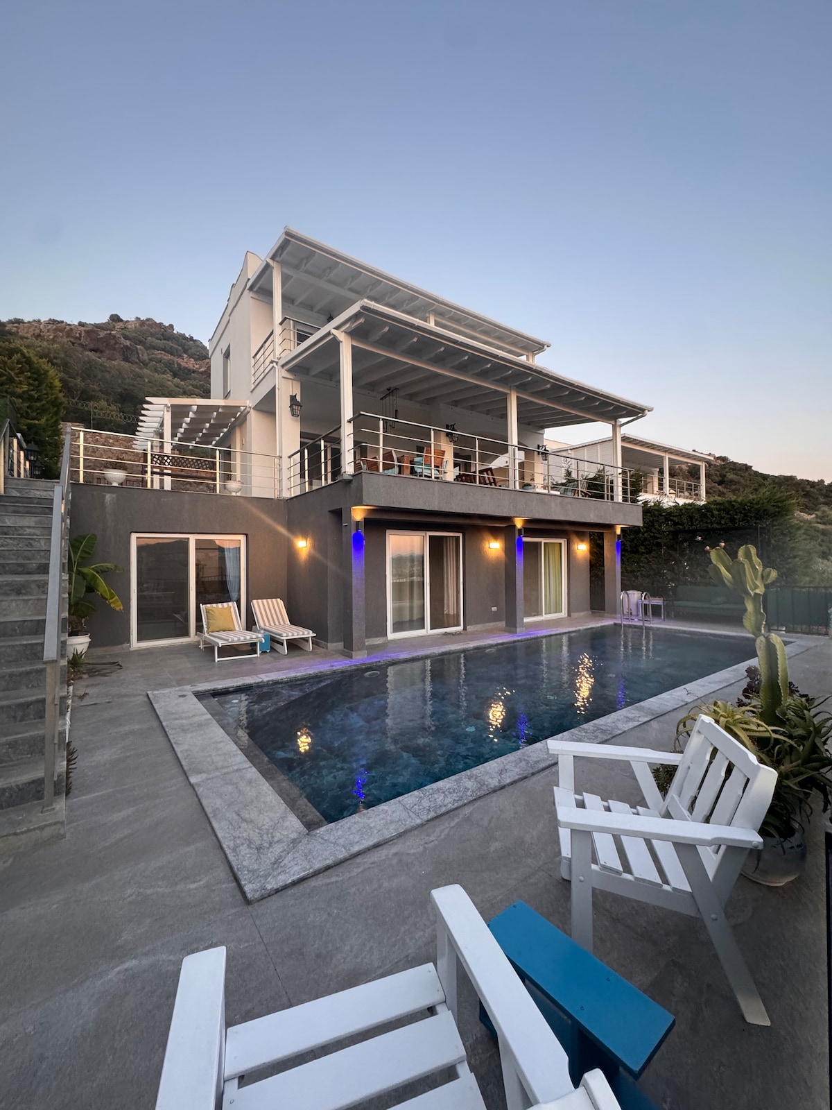 The exterior of the villa is shown at dusk, featuring a private swimming pool surrounded by seating and greenery. The spacious balcony above is illuminated, offering an inviting space for relaxation. The modern structure incorporates large glass doors for natural light.