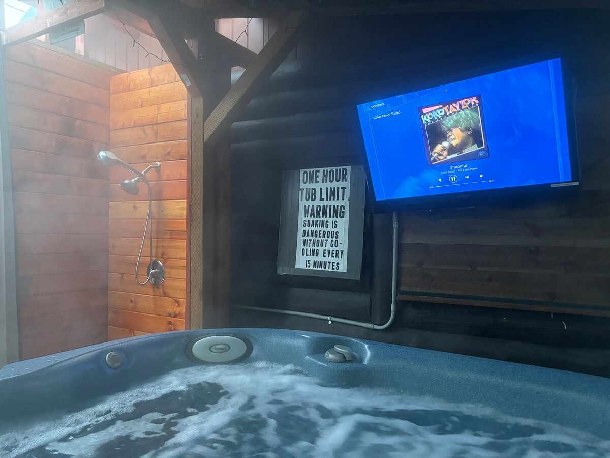 A hot tub is situated within a wooden enclosure, with wood walls providing a rustic atmosphere. A television is mounted on the wall, displaying content. A sign with safety instructions is visible nearby, and an outdoor shower is positioned beside the hot tub.