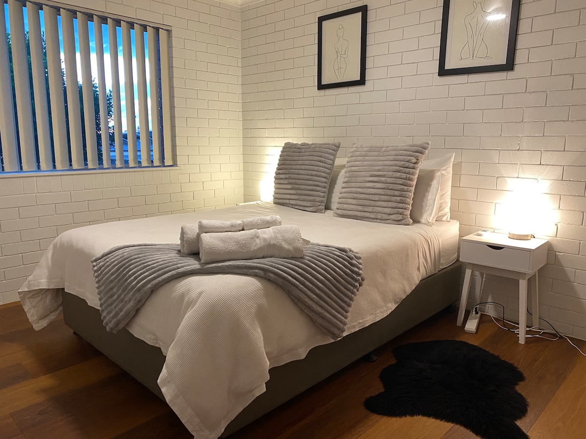 A spacious bedroom features a large bed with light bedding and decorative pillows. Soft lighting is provided by two bedside lamps placed on white nightstands. Neutral tones are complemented by a textured throw and a small rug, while natural light filters through vertical blinds.