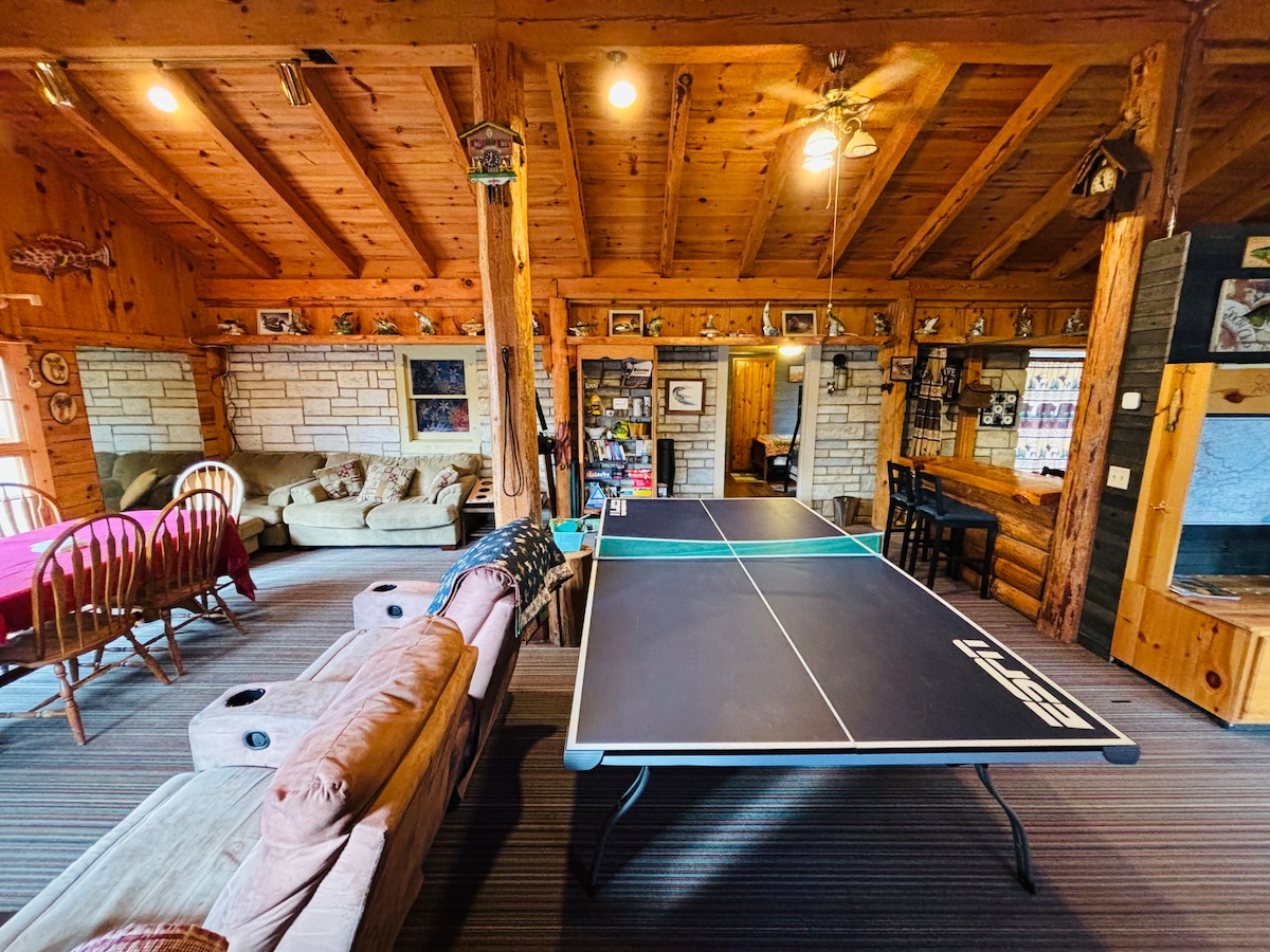 An inviting log cabin interior is presented, featuring a ping pong table at the center. Soft seating options are arranged around the room, and various decorative elements adorn the walls. A cozy dining area is visible in the background, contributing to a friendly gathering space.