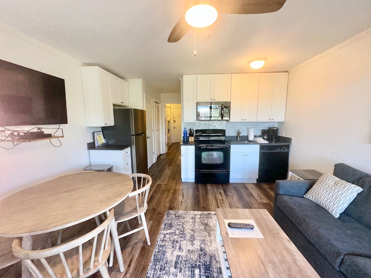 The open living space showcases a kitchen with modern appliances, including a refrigerator and oven. A dining table with two chairs is positioned beside a cozy sofa. Light wood flooring complements the neutral decor, enhancing the bright ambiance of the room.