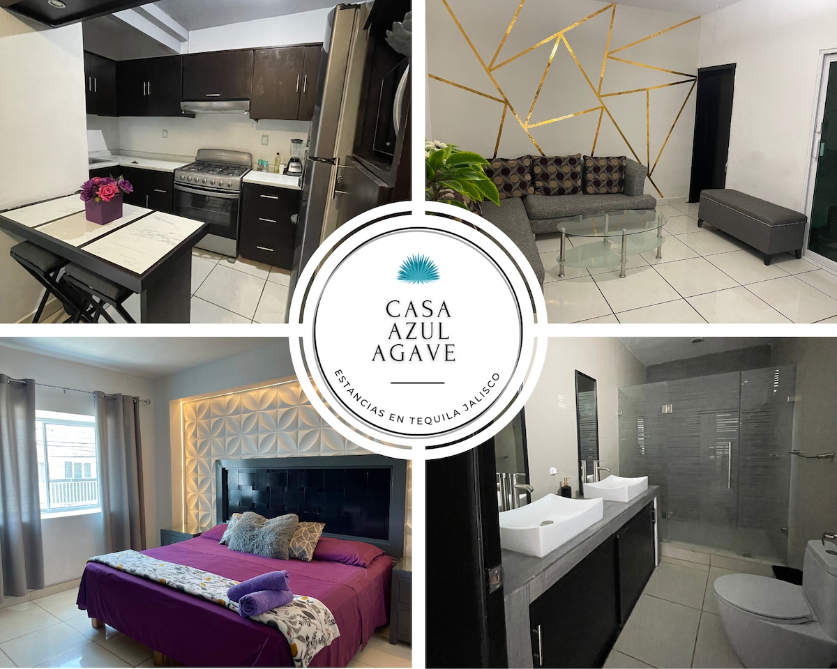 A modern kitchen includes black cabinetry and stainless-steel appliances, complemented by a dining area with a glass table. A comfortable seating area features a contemporary sofa and a coffee table. The main bedroom showcases a plush bed with dark linens and an elegant headboard, with a stylish ensuite bathroom visible.