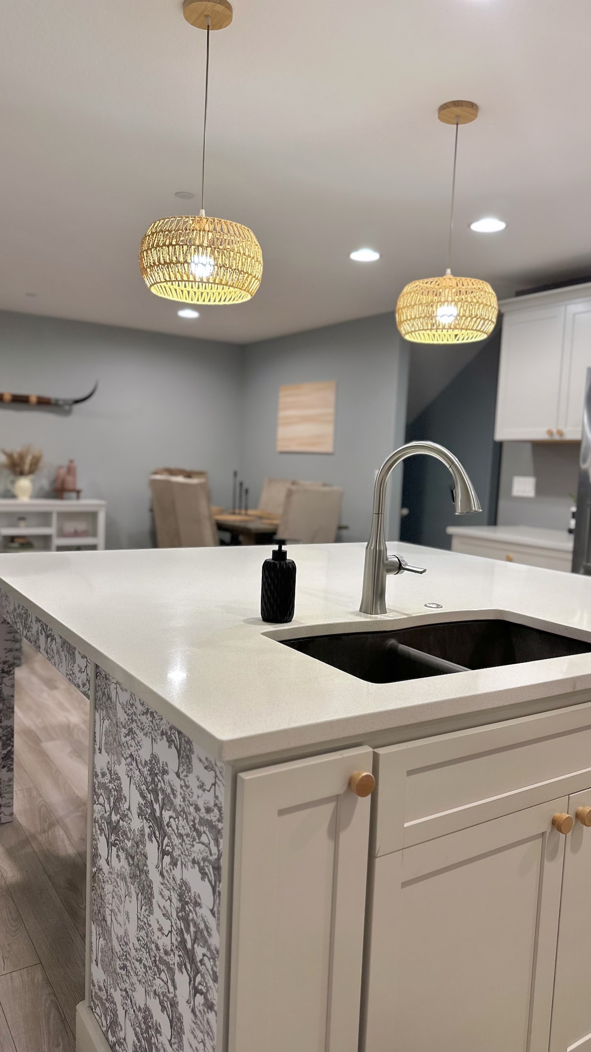 A modern kitchen is viewed, featuring a spacious island with a double sink. Elegant pendant lights hang overhead, illuminating the area. The light cabinetry complements the neutral walls, while dining chairs are seen in the background, enhancing the inviting atmosphere.