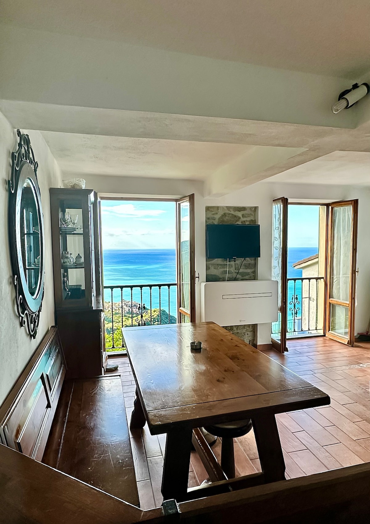 A dining area features a sturdy wooden table with benches, illuminated by natural light filtering through large glass doors. A display cabinet with decorative items is positioned against the wall, while a television is mounted nearby, offering views of the coastline beyond.
