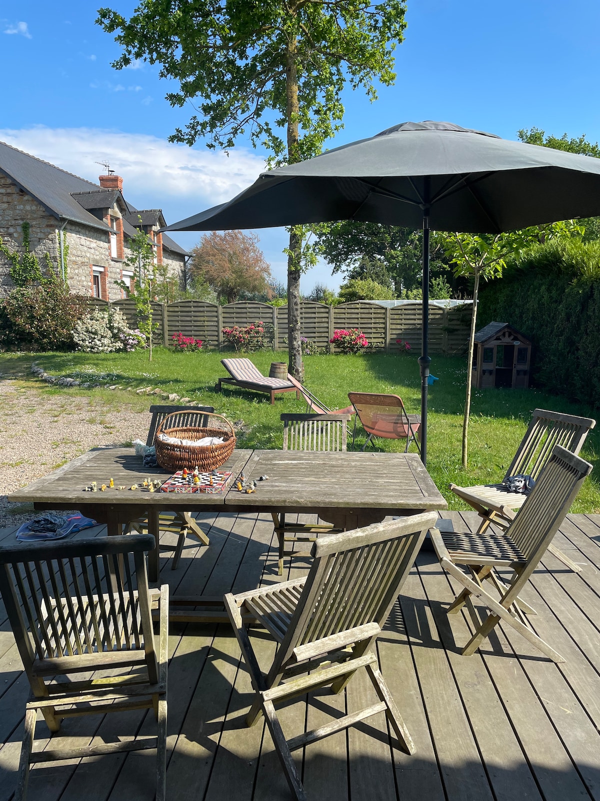 An outdoor terrace features a wooden table surrounded by six chairs, shaded by a large umbrella. Sun loungers are positioned on the grass, next to a fenced area adorned with blooming flowers. The serene garden is framed by hedges and trees.