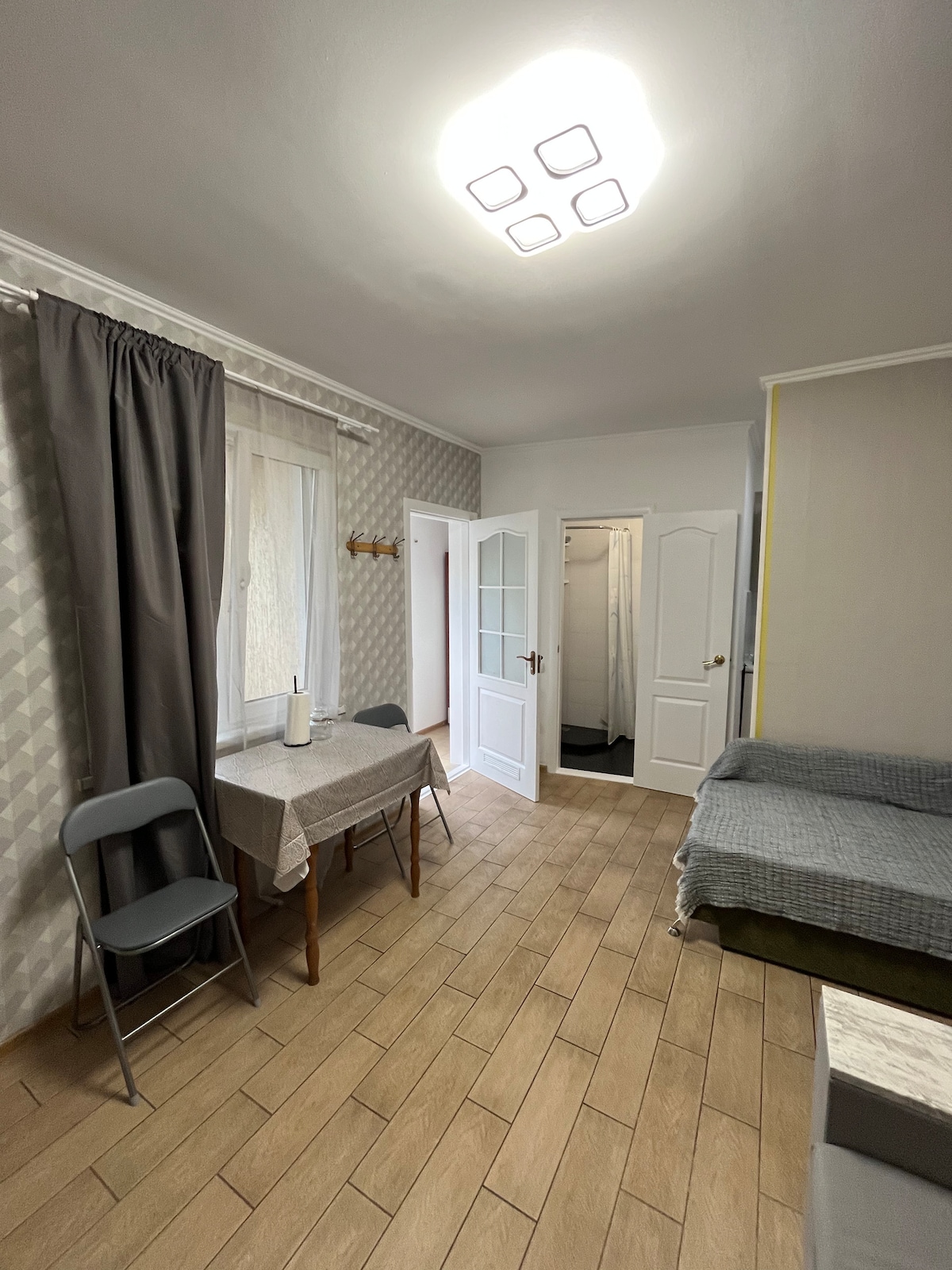 A small, inviting interior space is visible, featuring a table with two chairs beside a window dressed in light curtains. A cozy sleeping area is seen in the background, along with a separate area leading to a bathroom marked by white doors.