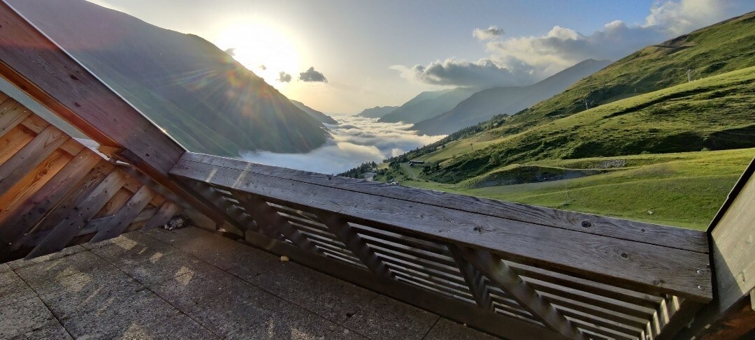A wooden balcony offers a stunning view of rolling hills and valleys bathed in soft morning light. The sunlight creates a serene atmosphere, illuminating the landscape below, which is occasionally shrouded in mist, enhancing the sense of tranquility.