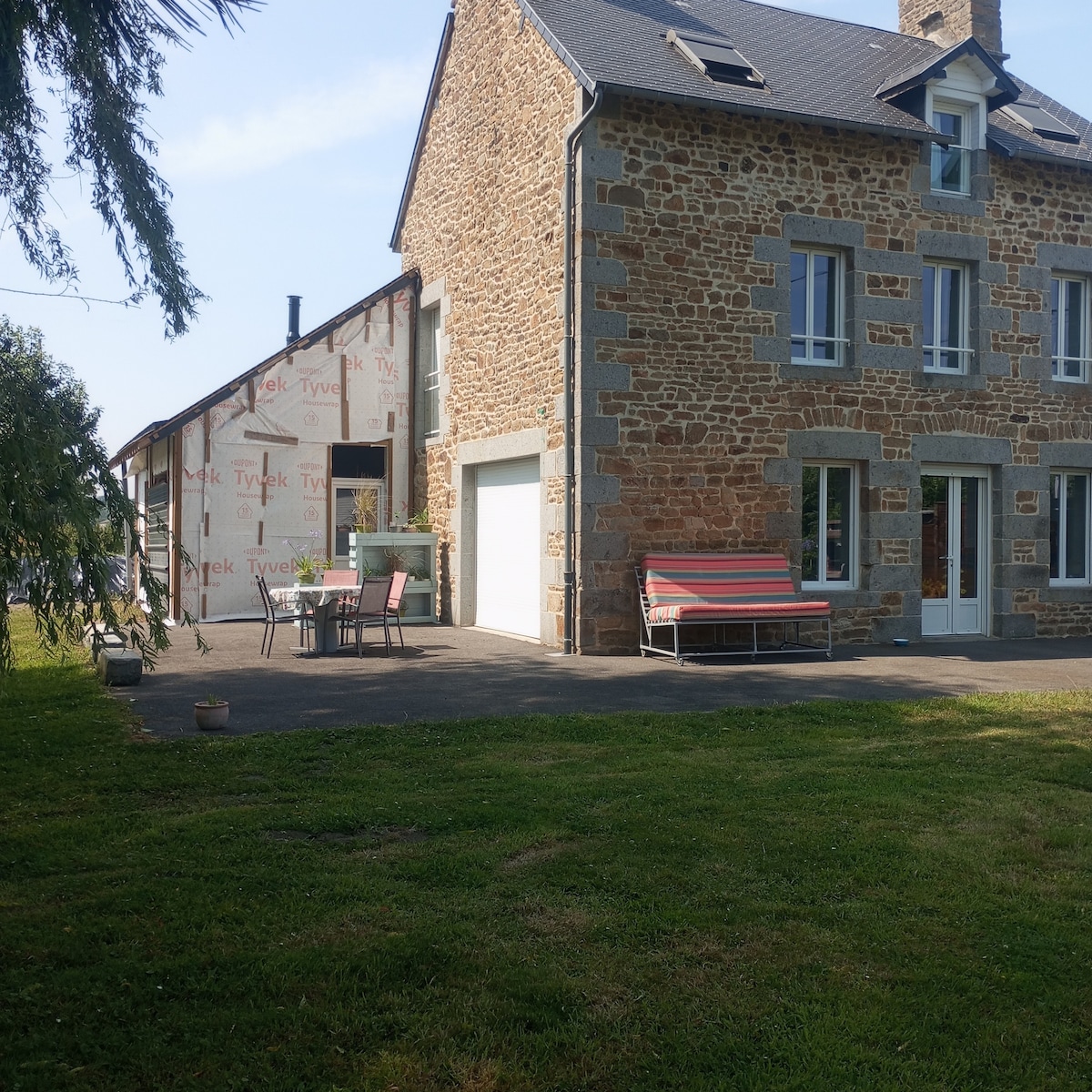 A charming villa is shown, featuring a stone facade and a spacious terrace that includes outdoor seating. The area is surrounded by green grass and trees, creating a serene environment, while large windows provide ample natural light to the interior.