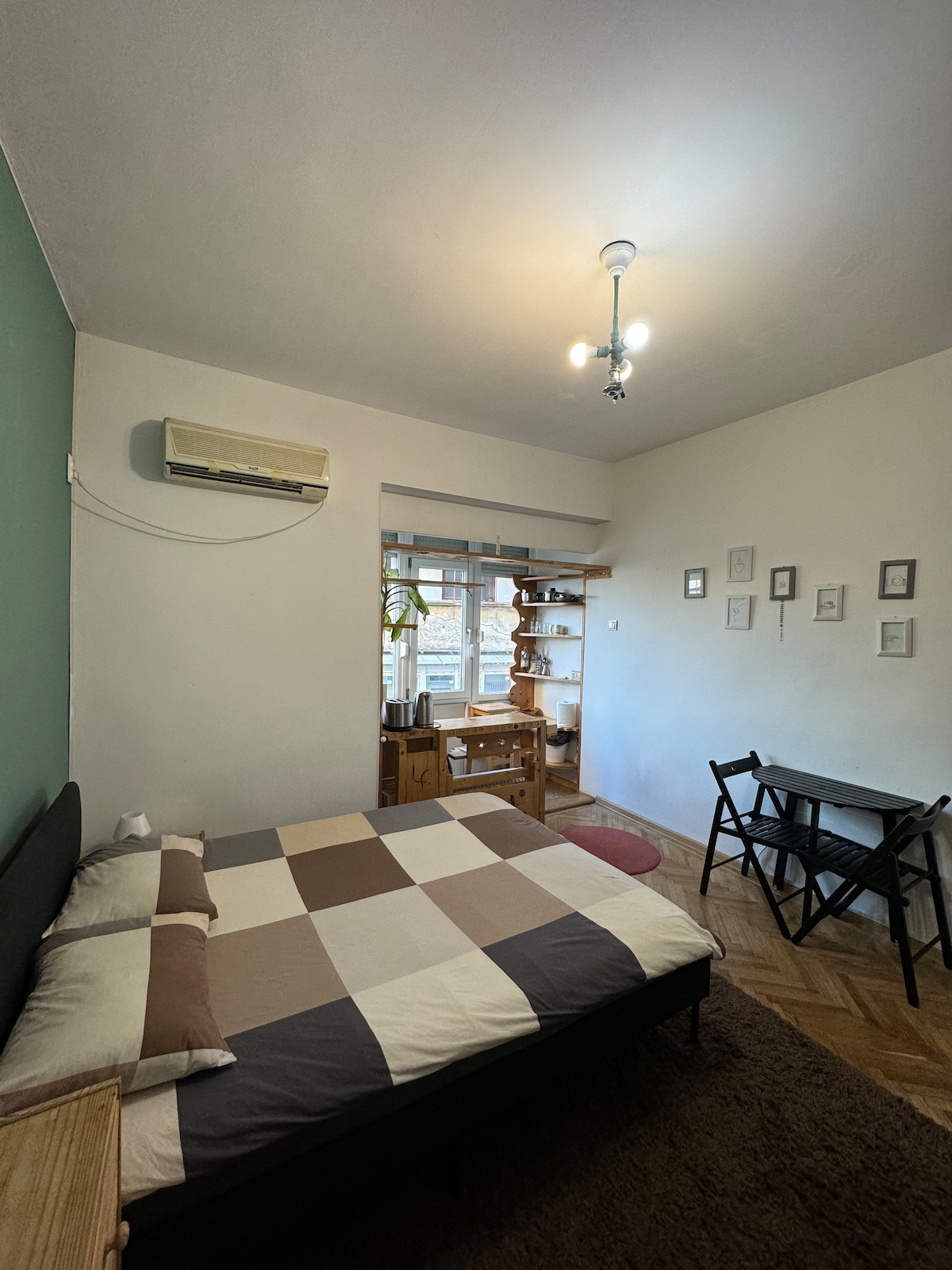 The studio features a spacious bed with a patterned blanket, complemented by a soft rug. Natural light enters through large windows. A small dining area with two chairs is positioned next to a wooden shelving unit, housing decorative items and plants.