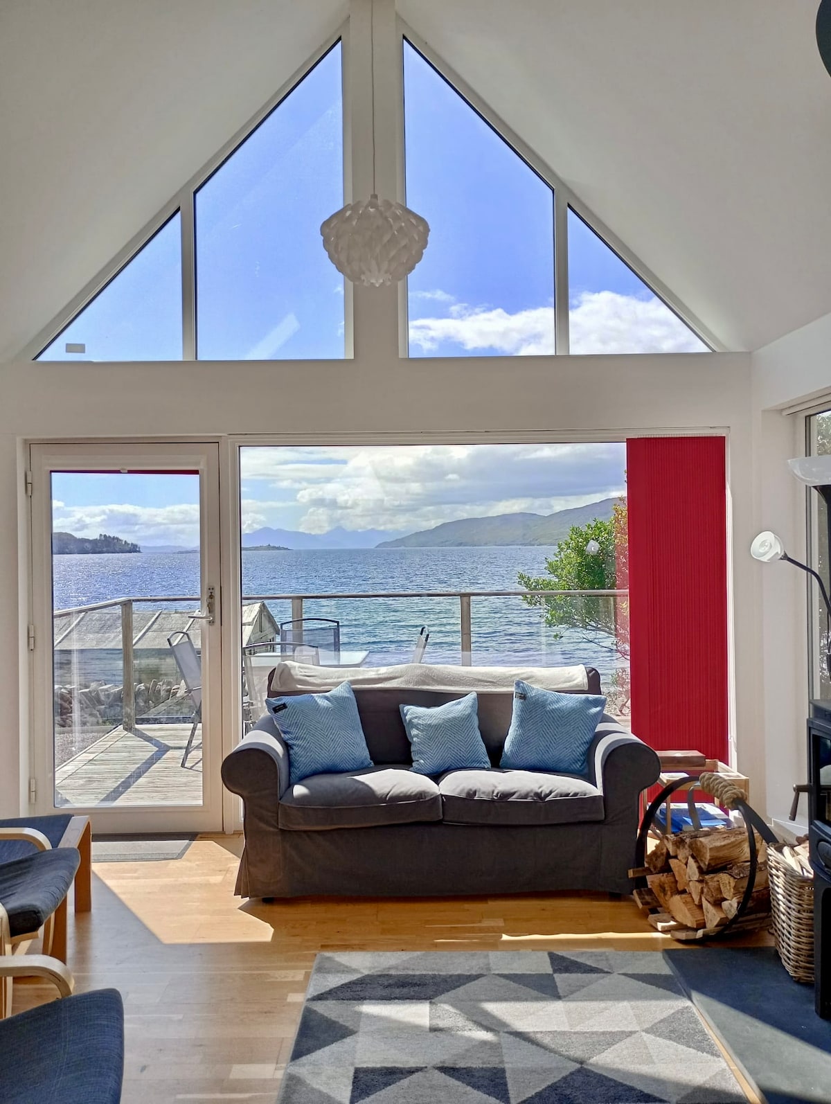 A bright living room features a gray sofa with blue cushions, positioned in front of large triangular windows that offer expansive views of the water. A sliding glass door leads to a deck, and a small coffee table rests on a patterned rug. Sunlight illuminates the space.