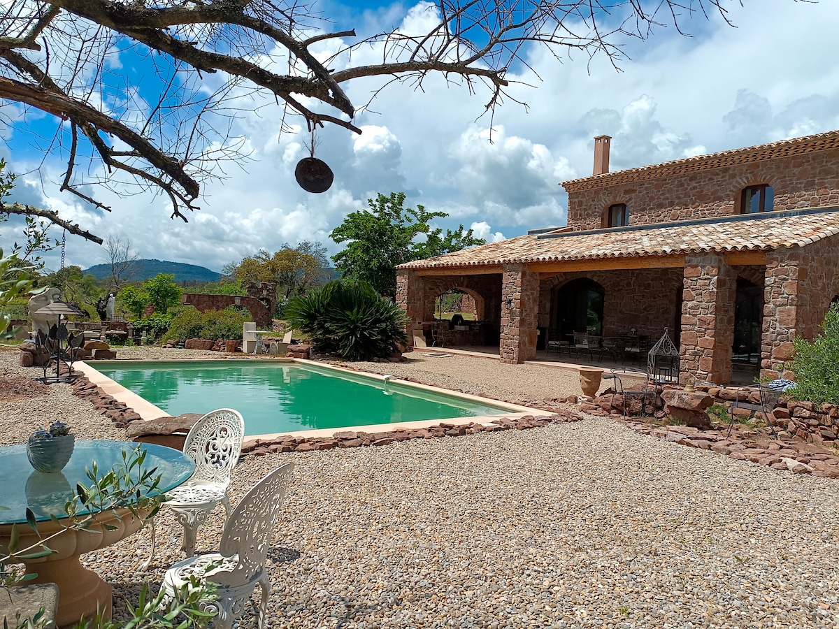 A stone house is situated beside a green swimming pool, surrounded by a gravel terrace. Lush vegetation frames the scene, with trees and plants adding to the natural ambiance. The sky is partly cloudy, providing a serene backdrop for the outdoor space.
