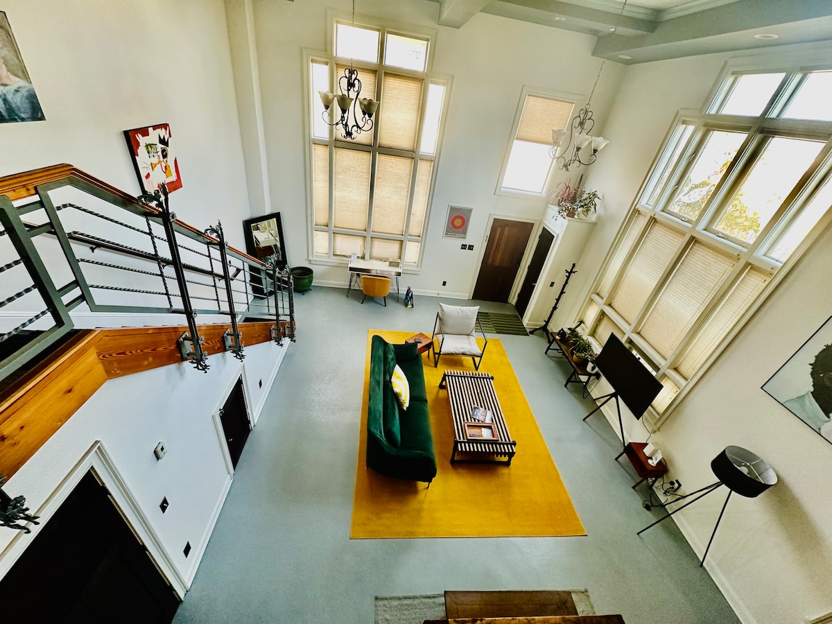 The Great Room features high ceilings with multiple large windows providing ample natural light. A green couch and an accent chair are positioned around a striped coffee table on a vibrant yellow area rug. A staircase leads to the mezzanine, adding architectural interest.