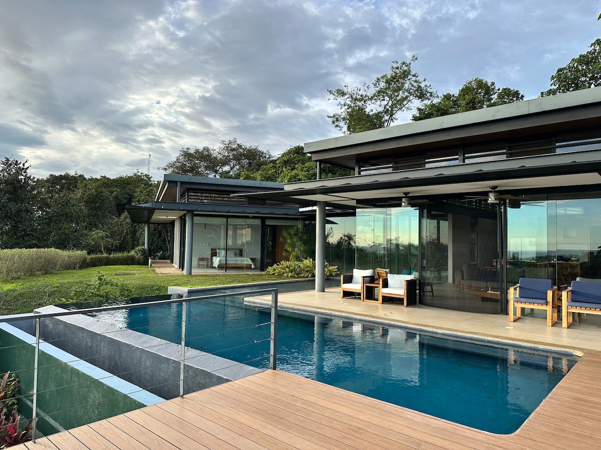 A modern glass house is set against a lush landscape, featuring a private pool with a sleek design and wooden decking. Comfortable outdoor seating is positioned nearby, allowing for relaxation while enjoying the scenic views of the surrounding nature preserve.