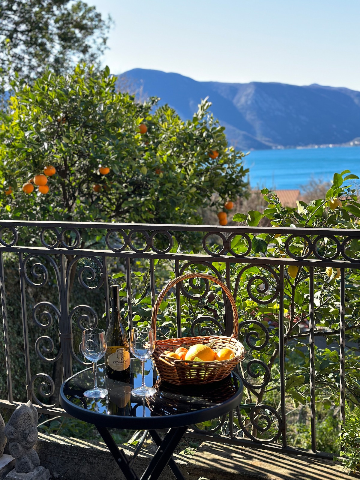 A balcony table is set with a woven basket filled with oranges and two glasses beside a bottle. A verdant garden features orange trees, with distant mountains and the sea visible in the background, creating a serene outdoor setting.