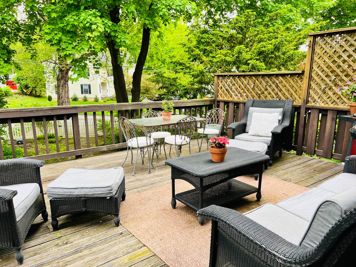 An outdoor deck area features a seating arrangement with a dark wicker coffee table, surrounded by four metal chairs and a small potted plant. Two lounge chairs are positioned nearby for relaxation, while greenery provides a natural backdrop to the space.