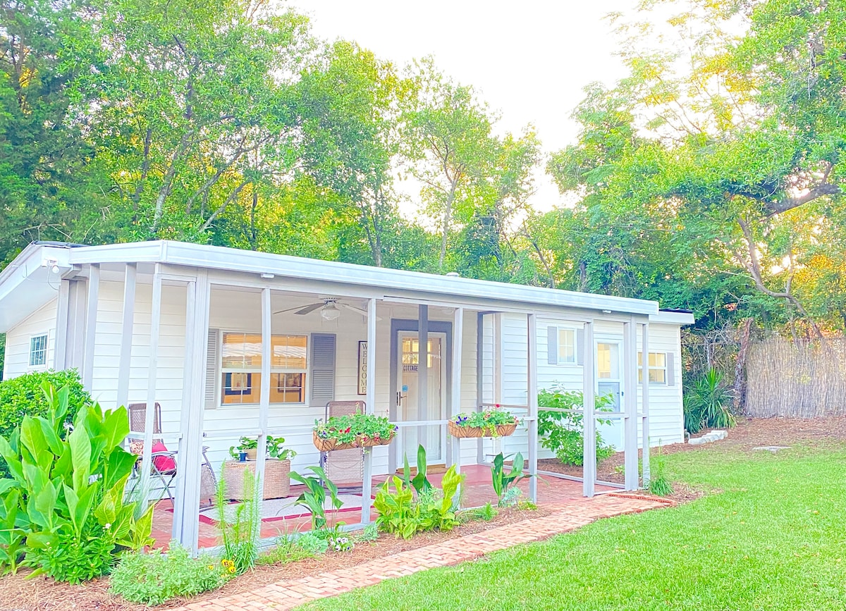 A cottage exterior is shown, featuring a welcoming porch with seating areas surrounded by lush greenery. Planters with vibrant foliage line the porch, while natural light bathes the space, creating a serene atmosphere among the surrounding trees.