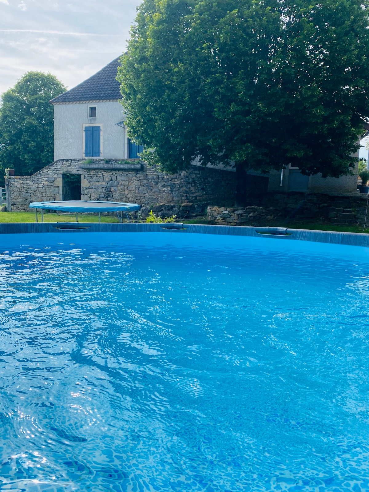 A clear, inviting swimming pool reflects sunlight, surrounded by well-maintained greenery. In the background, a historical stone building with a single window is partially visible, complemented by a large tree providing shade.