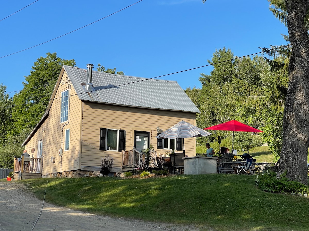 A charming cottage is set against a clear blue sky, featuring a spacious patio with umbrellas for outdoor relaxation. The light-colored exterior contrasts with the greenery surrounding the property, while a gravel driveway leads to the inviting entrance.