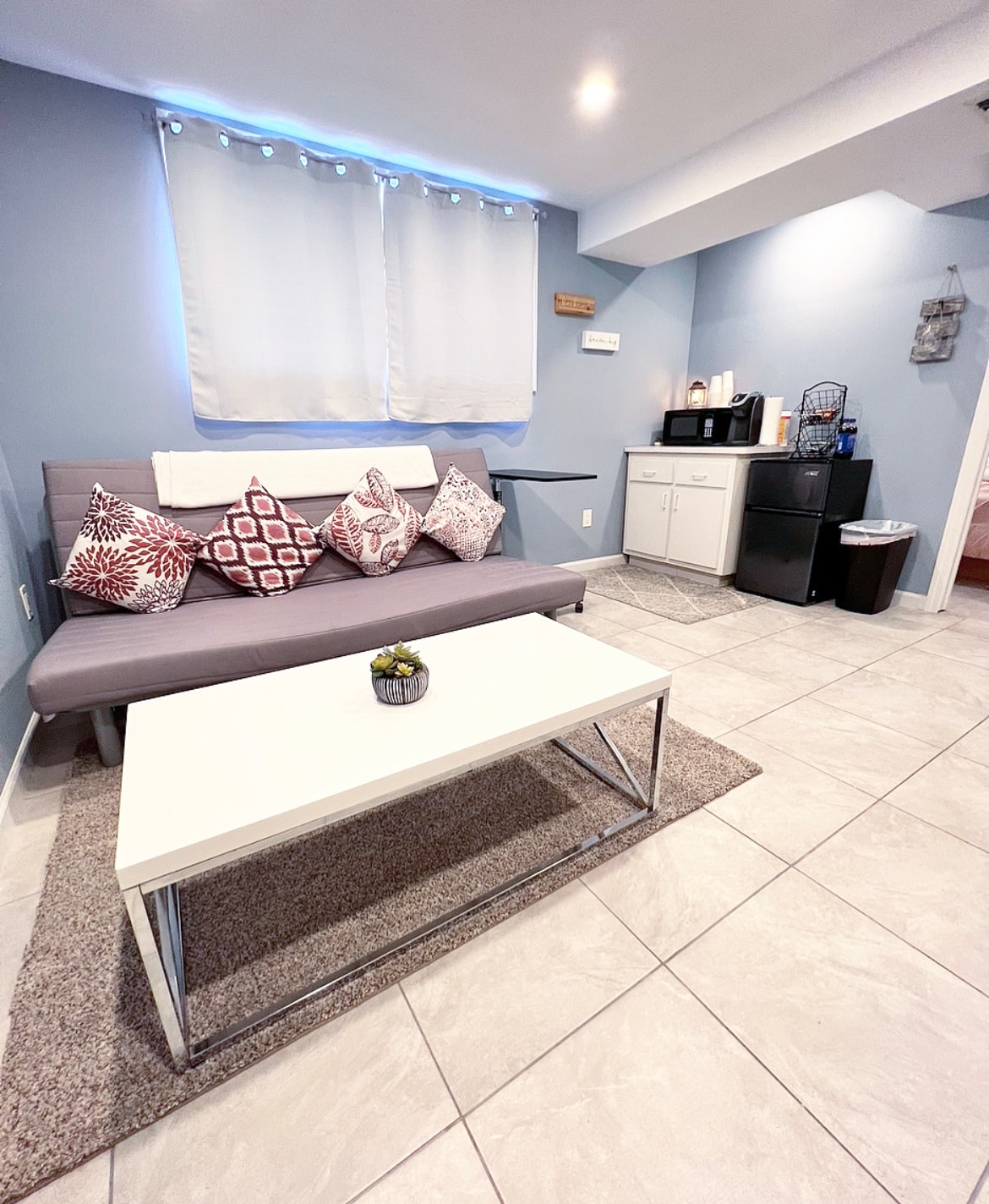 A modern living area is showcased with a gray futon adorned with decorative pillows. A white coffee table sits on a soft area rug, while a microwave and refrigerator are positioned against the wall. Soft lighting enhances the clean tile flooring.
