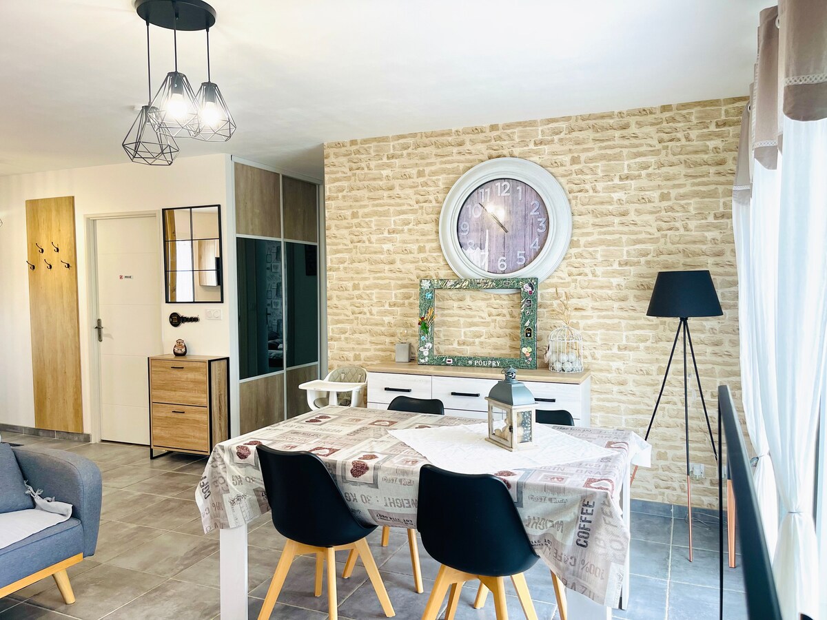 A dining area is depicted with a modern white table surrounded by four black chairs. A large clock is mounted on a textured brick wall, complementing a stylish sideboard and decorative mirror. Natural light floods in from nearby windows adorned with sheer curtains.
