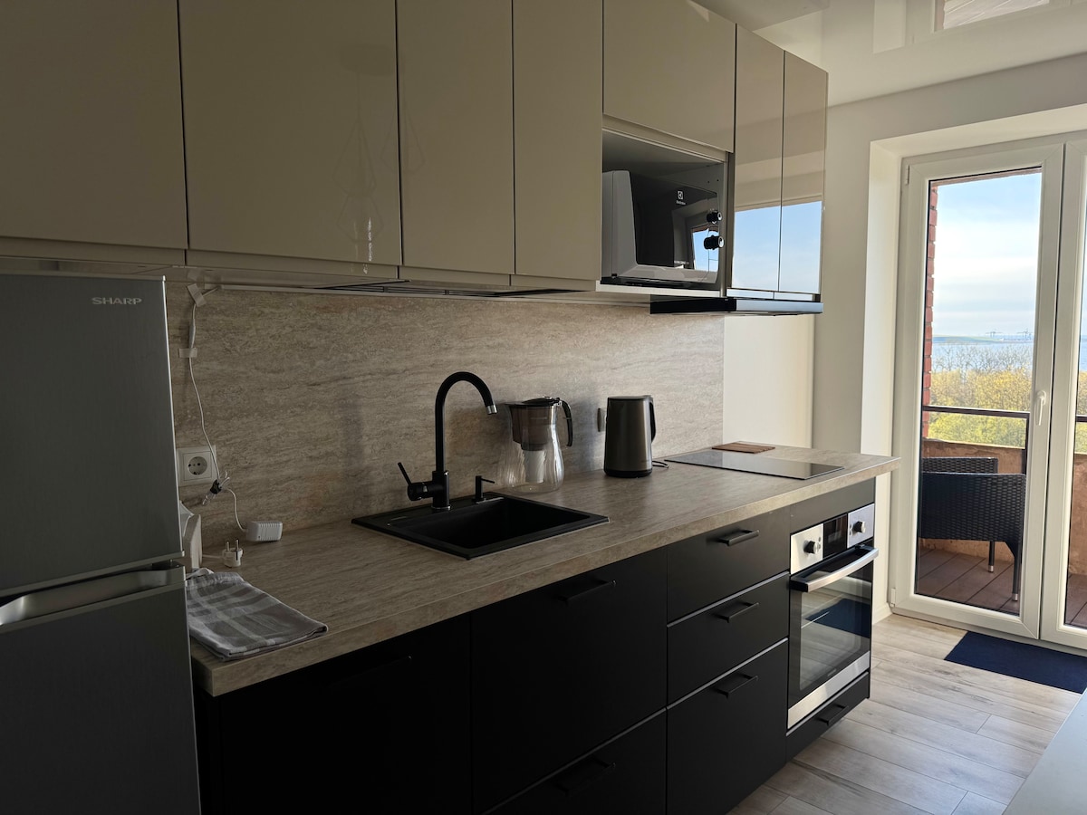 A modern kitchen features sleek black and beige cabinetry with minimalistic design. Stainless steel appliances, including a refrigerator and an oven, are integrated into the space. A window allows natural light to illuminate the countertop, creating a functional cooking area.