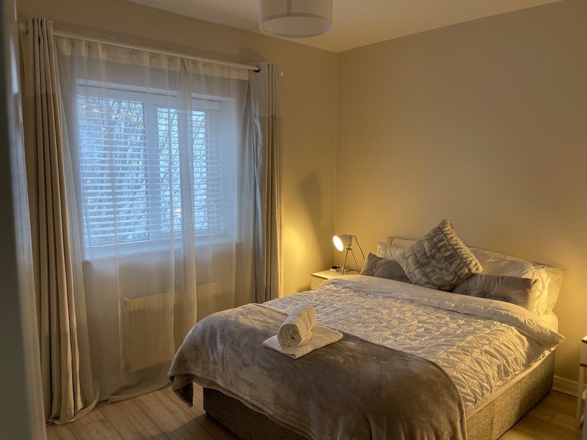 A softly lit bedroom is equipped with a spacious bed dressed in light textiles. The room features a large window draped with sheer curtains, allowing gentle light to filter in. A bedside lamp and a neatly placed towel complete the inviting space.