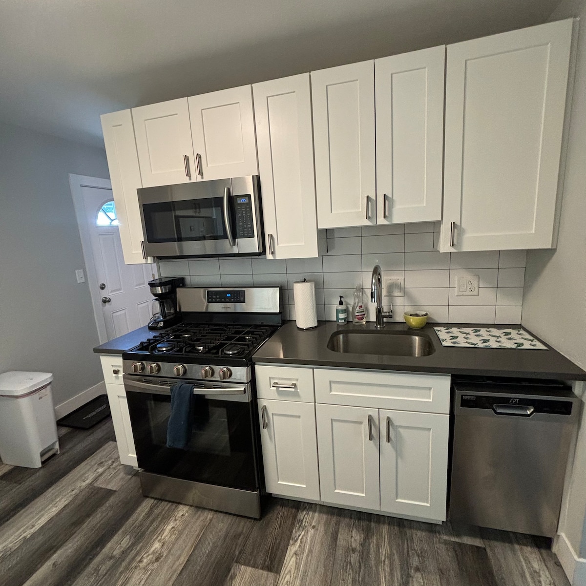The kitchen features modern white cabinetry and stainless steel appliances, including a gas stove and dishwasher. A sleek countertop provides ample workspace, with a coffee maker and sink visible. The door leads to the entry, and a light-colored floor complements the overall design.
