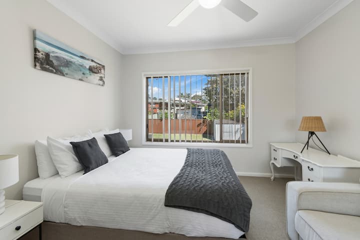 A bright and calming second bedroom located downstairs, featuring a comfortable queen bed, soft coastal touches, a sofa, and a small vanity desk for getting ready.