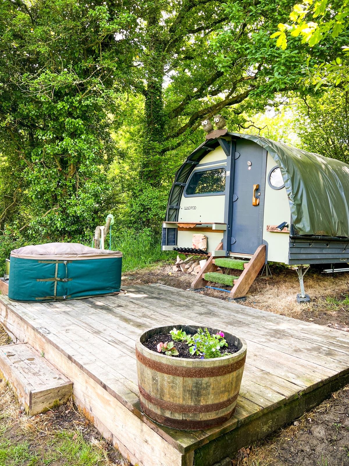 A cozy Landpod is nestled among greenery, featuring a covered entrance and a small seating area. Nearby, a wooden deck holds a circular hot tub and a barrel planter with colorful flowers, harmonizing with the natural surroundings.