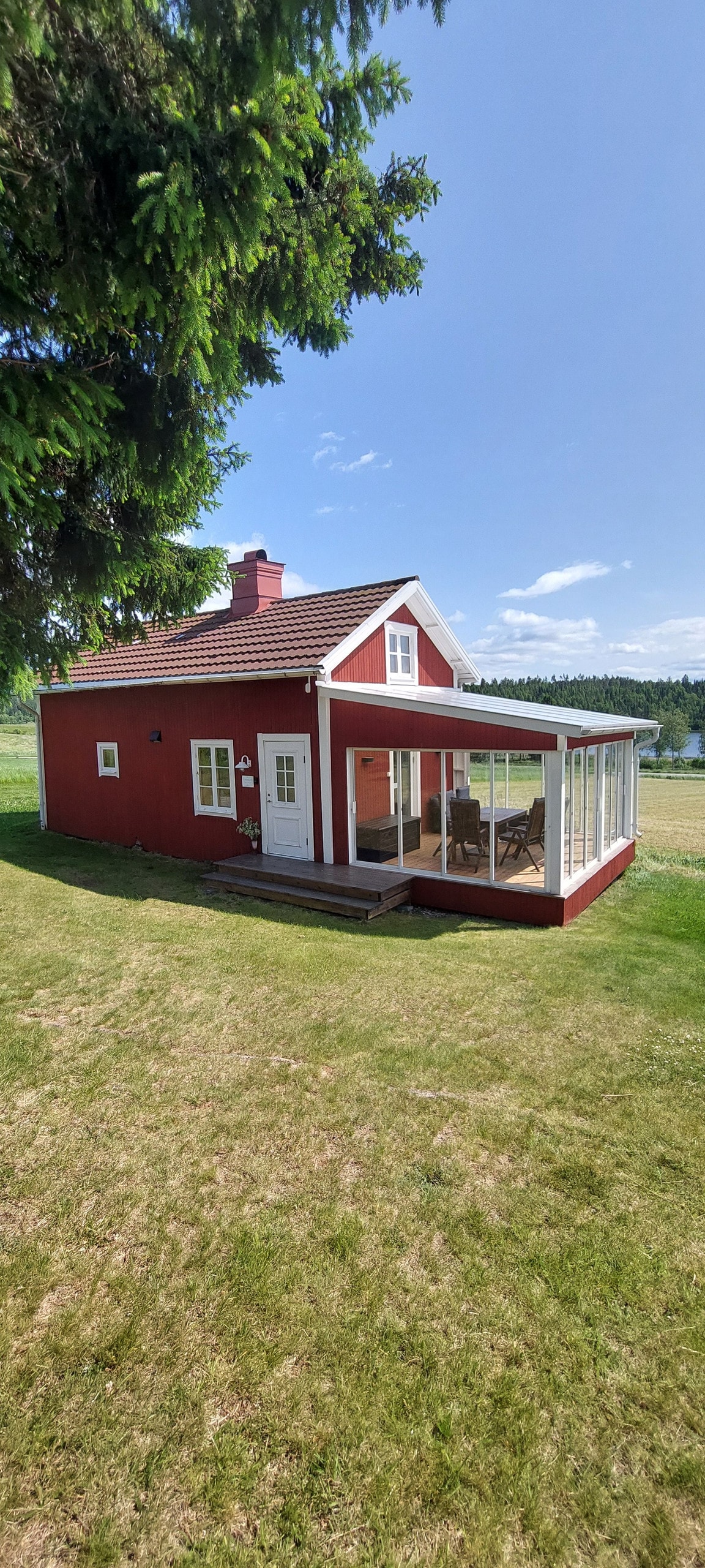 A red cottage with a white porch features a spacious, enclosed veranda adorned with outdoor furniture. The surrounding grassy area is open, offering a clear view of the landscape. The structure showcases a classic Swedish design, set against a backdrop of blue skies.