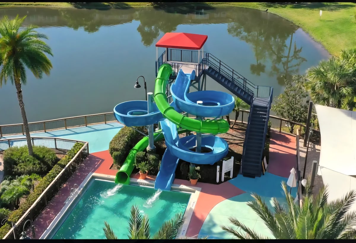 A vibrant water slide complex is featured alongside a sparkling pool, surrounded by tropical landscaping. The structure includes dual twisting slides in shades of green and blue, with a staircase leading to the landing deck. A serene lake is visible in the background.