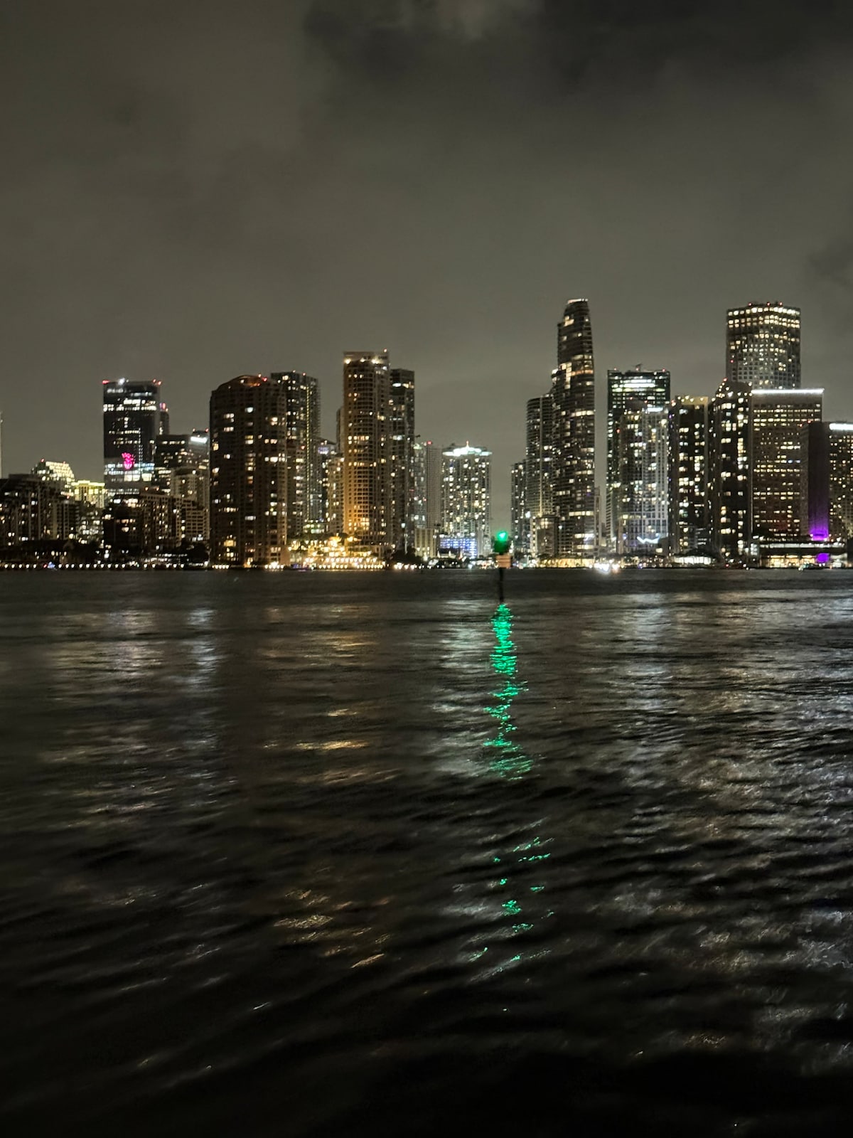 A vibrant city skyline is illuminated at night, with numerous high-rise buildings reflecting colorful lights on the water. The scene includes a prominent green light, enhancing the dynamic atmosphere. The surrounding water appears calm, creating a striking contrast with the bustling city view.