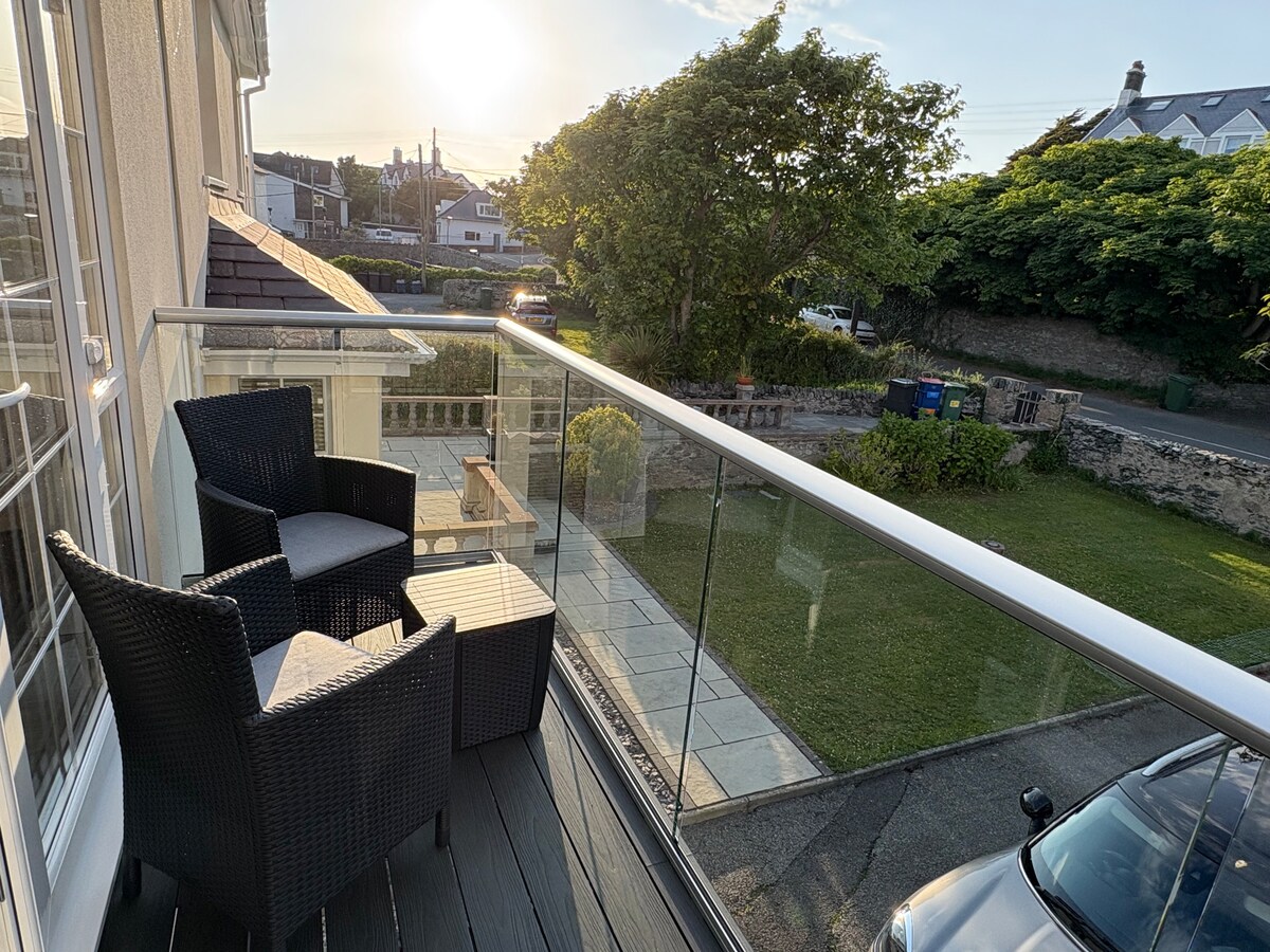 A balcony area is showcased, furnished with two comfortable chairs and a small table. Clear glass balustrades provide unobstructed views of the surrounding greenery and quiet road. The setting is bathed in soft natural light from the setting sun, enhancing the tranquil atmosphere.
