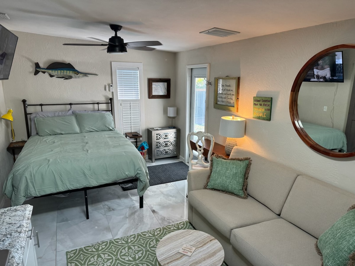 An efficiency space features a queen bed with light green bedding, complemented by a comfortable pull-out couch. A round mirror reflects the soft lighting and decorative elements. A small kitchenette area includes modern appliances, while a door leads to an outdoor space, allowing natural light to enter.