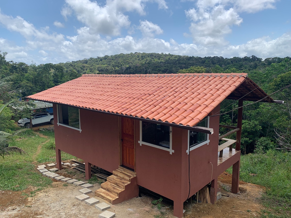 A rustic bungalow with a terracotta-tiled roof stands amidst lush greenery. Clear blue skies with soft clouds create a serene backdrop. The structure features wooden steps leading to a welcoming entrance, with large windows that enhance the connection with nature.